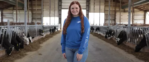 Woman standings in dairy barn wearing blue KUHN longs leeve shirt