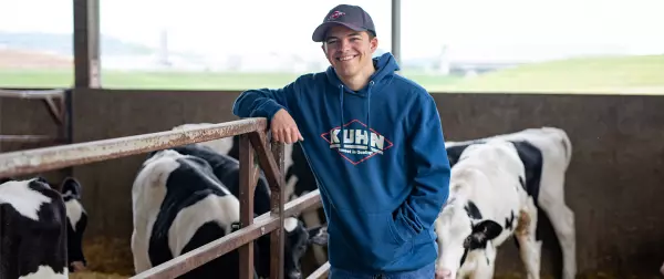 Young man stands in front of dairy calves wearing blue KUHN sweatshirt and KUHN hat